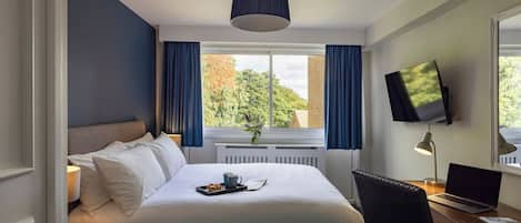 Ett hotellrum med en säng, ett skrivbord med en bärbar dator, en tv på väggen och ett fönster med gardiner.