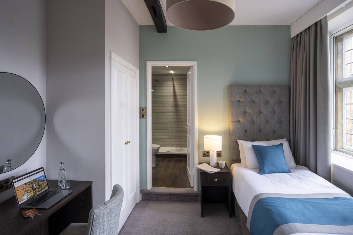 Une chambre d’hôtel équipée d’un lit, d’une table de chevet, d’une lampe et d’un bureau avec un ordinateur portable et une bouteille d’eau.