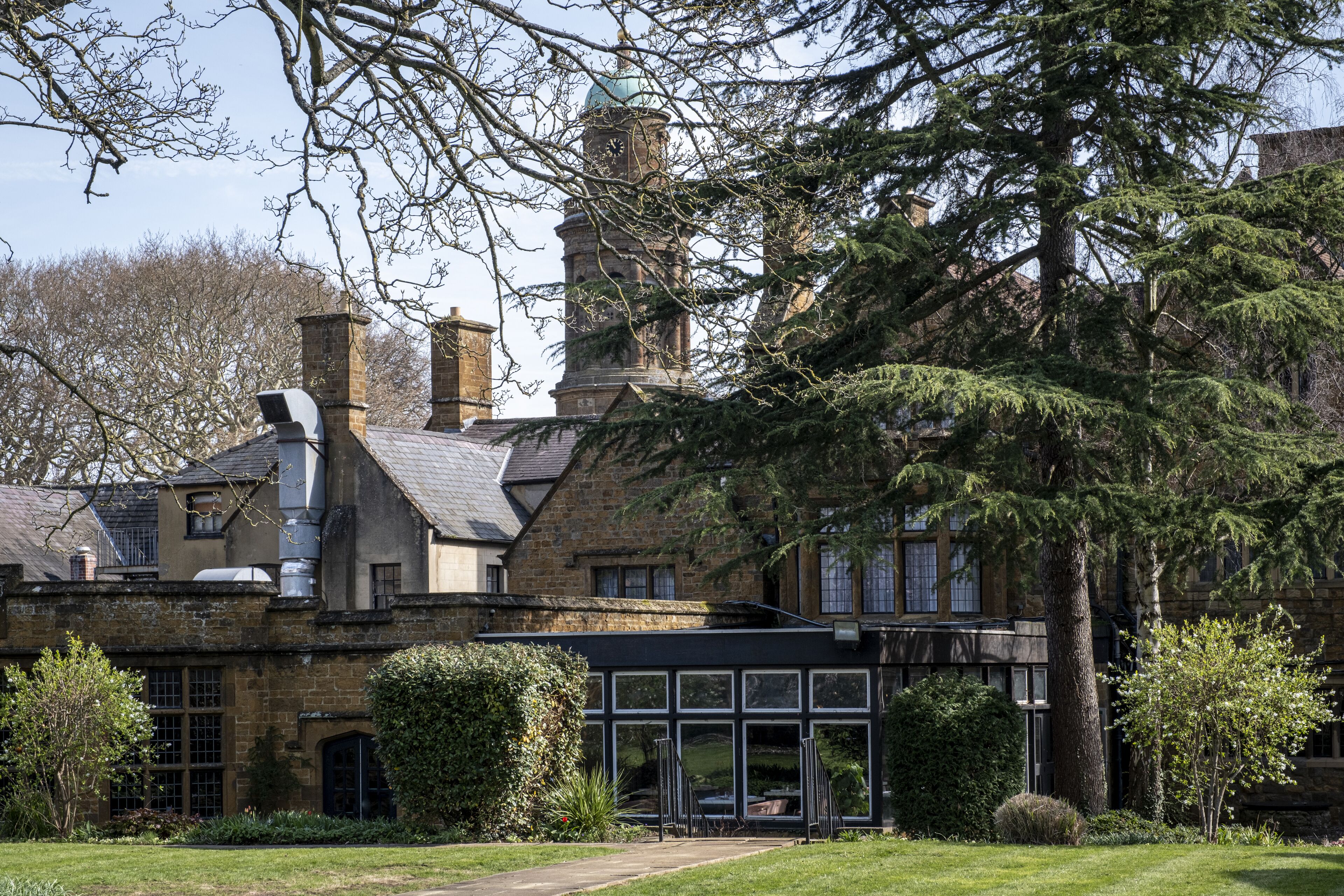 A historic stone building with a prominent clock tower, large windows, and a well-maintained garden.