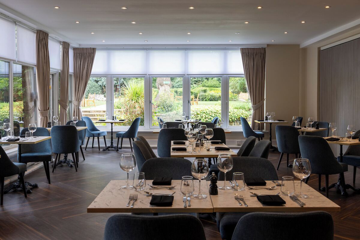 A restaurant with multiple tables and chairs, large windows with curtains, and a view of greenery outside.