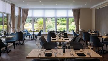 A restaurant with multiple tables and chairs, large windows with curtains, and a view of greenery outside.