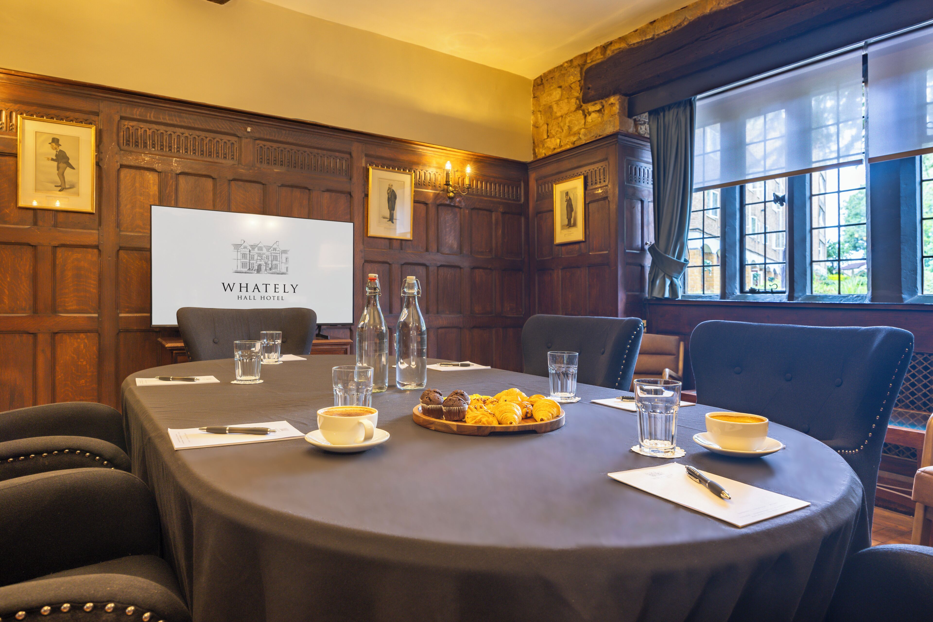 A conference room with a round table, chairs, a projector screen displaying 'WHATELY HALL HOTEL', and a window with a view of buildings.