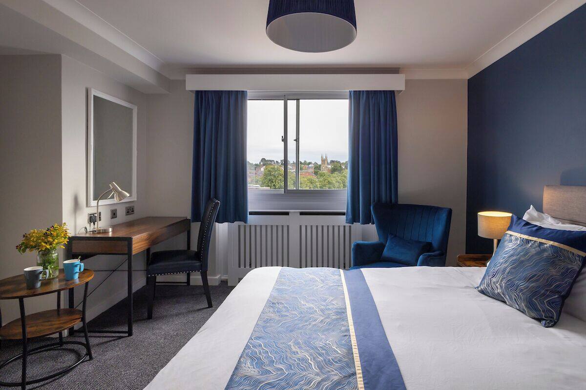 Une chambre d’hôtel avec un grand lit, un bureau avec une chaise, une fenêtre avec des rideaux et une vue sur le paysage urbain.