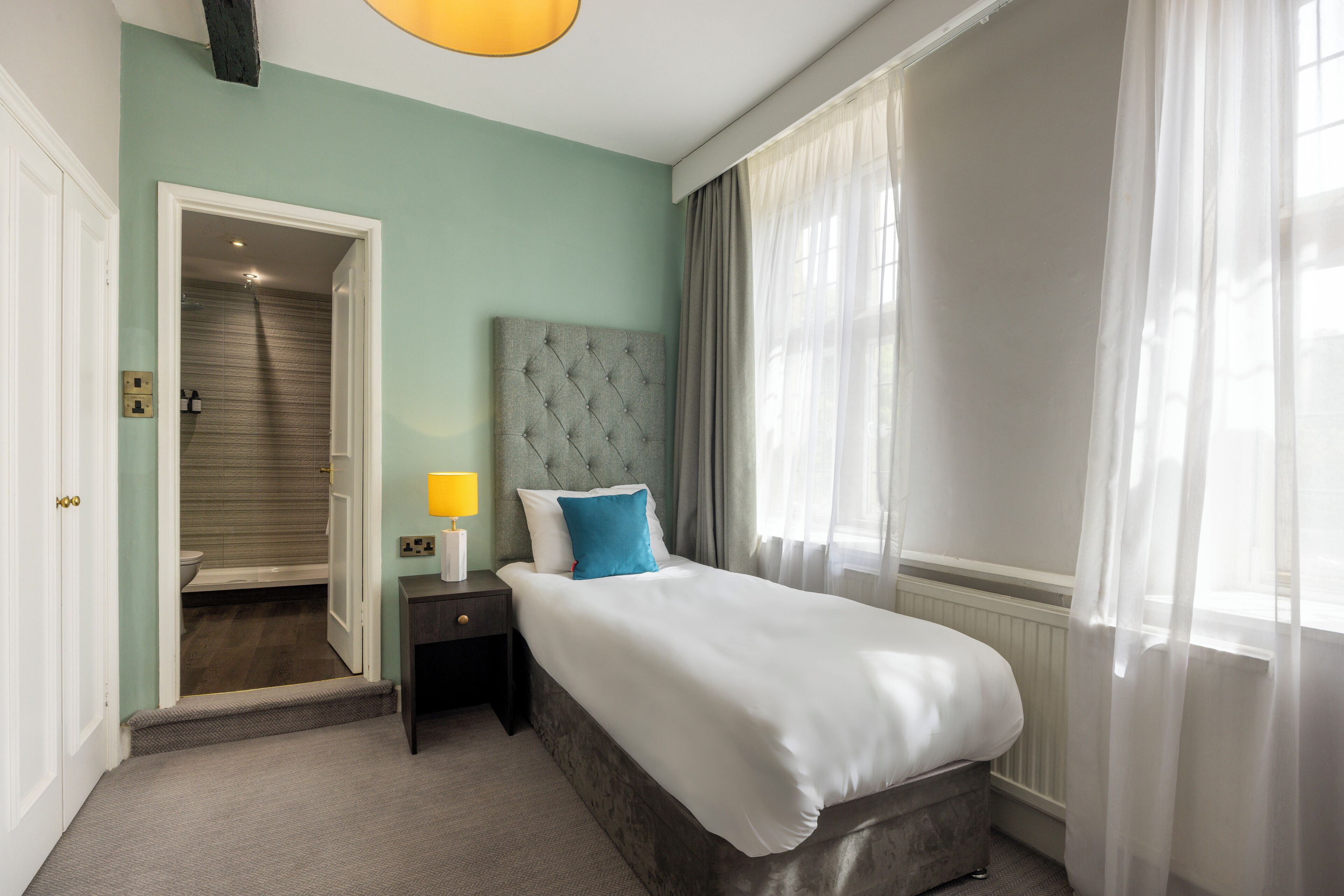A hotel room with a bed, a nightstand, a lamp, a bathroom door, and a window with curtains.