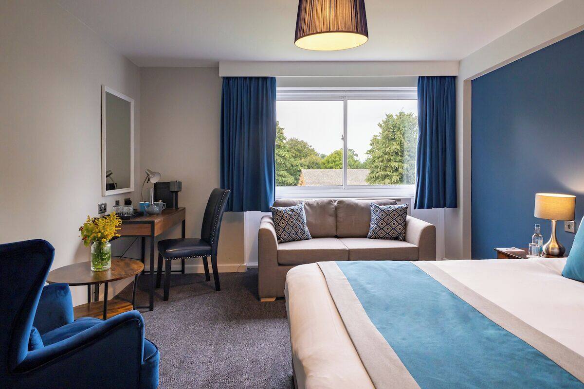 Une chambre d’hôtel avec un lit, un canapé, un bureau et une chaise. Il y a une fenêtre avec des rideaux, une lampe et un vase rempli de fleurs.