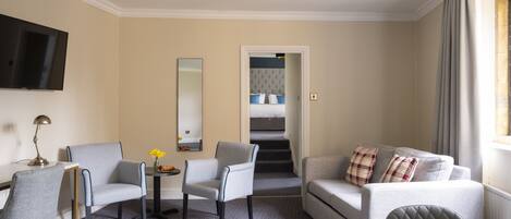 Habitación de hotel con sofá, dos sillones, una mesita con flores, televisión y espejo.