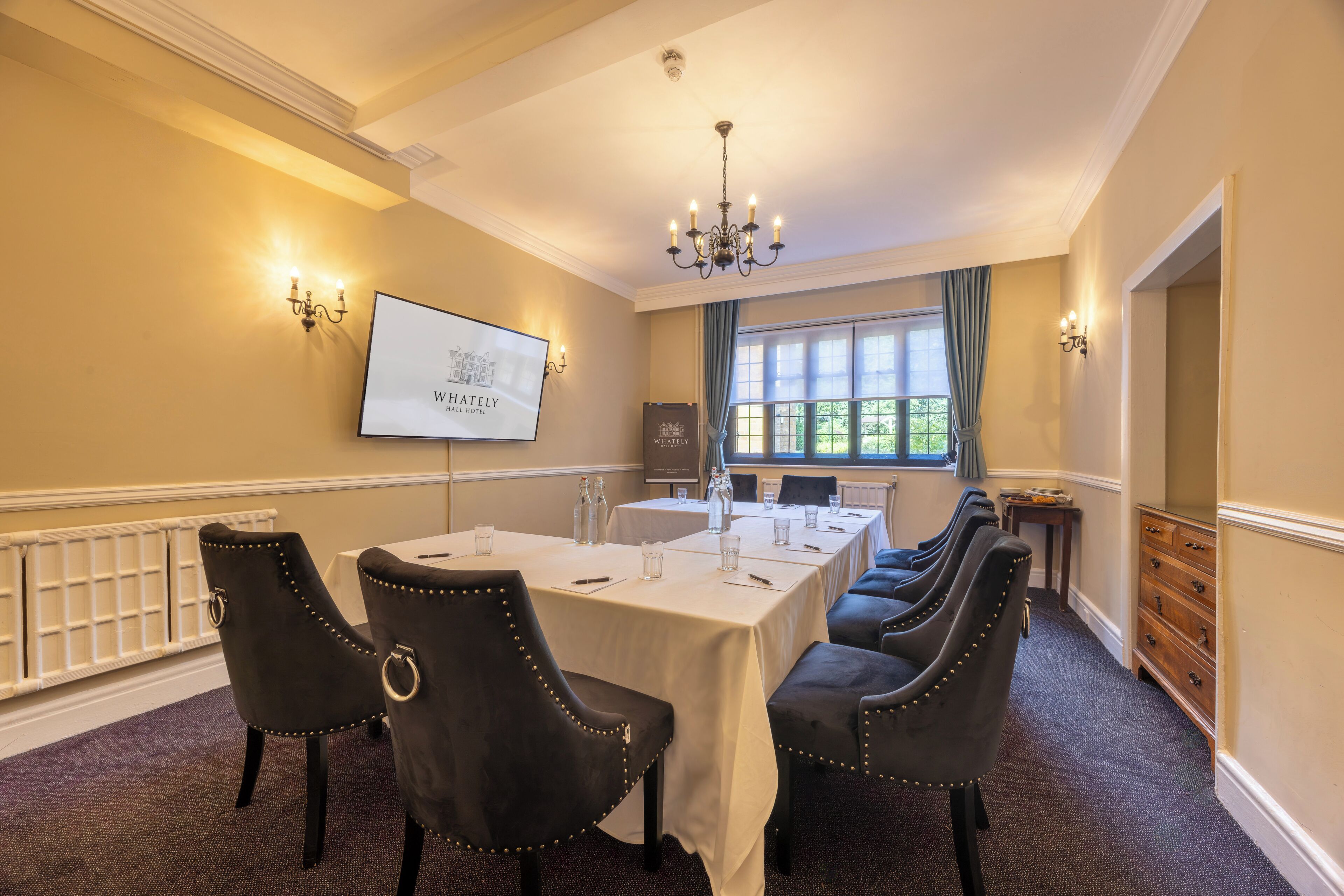 A conference room with a long table, black upholstered chairs, a projector screen, and a chandelier.