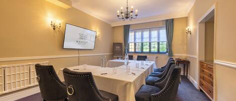 A conference room with a long table, black upholstered chairs, a projector screen, and a chandelier.
