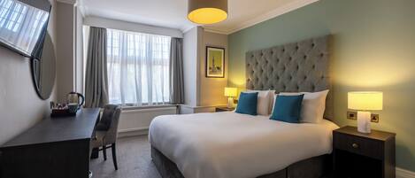 Habitación de hotel con una cama grande, un escritorio, una silla, una mesita de noche con lámpara y una ventana con cortinas.
