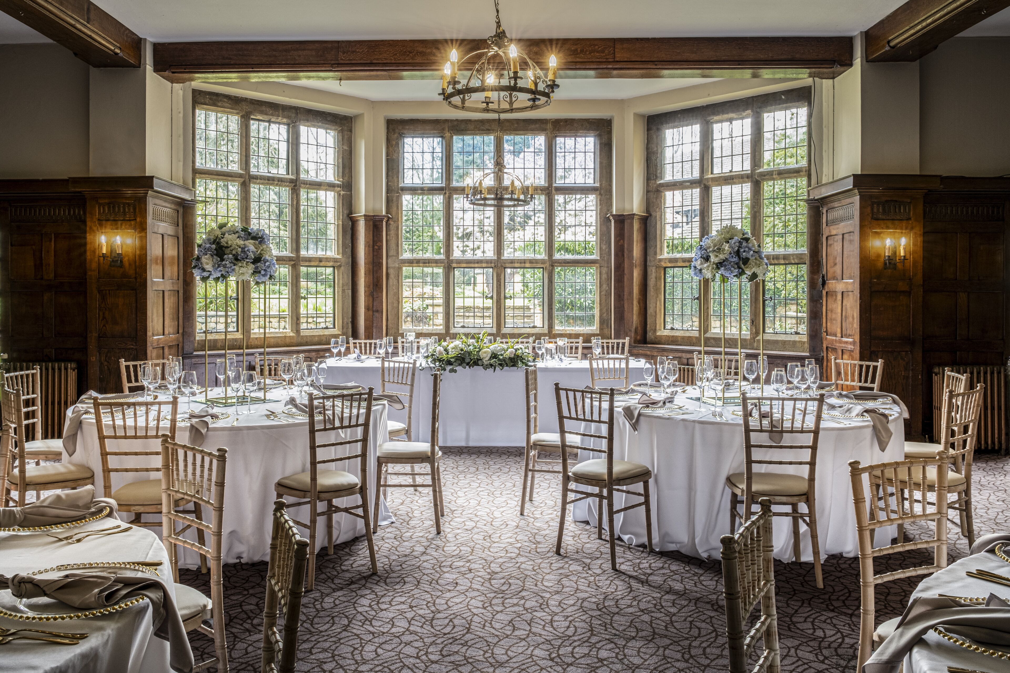 A formal dining room with multiple round tables set for a meal, large windows with a view, and a chandelier.
