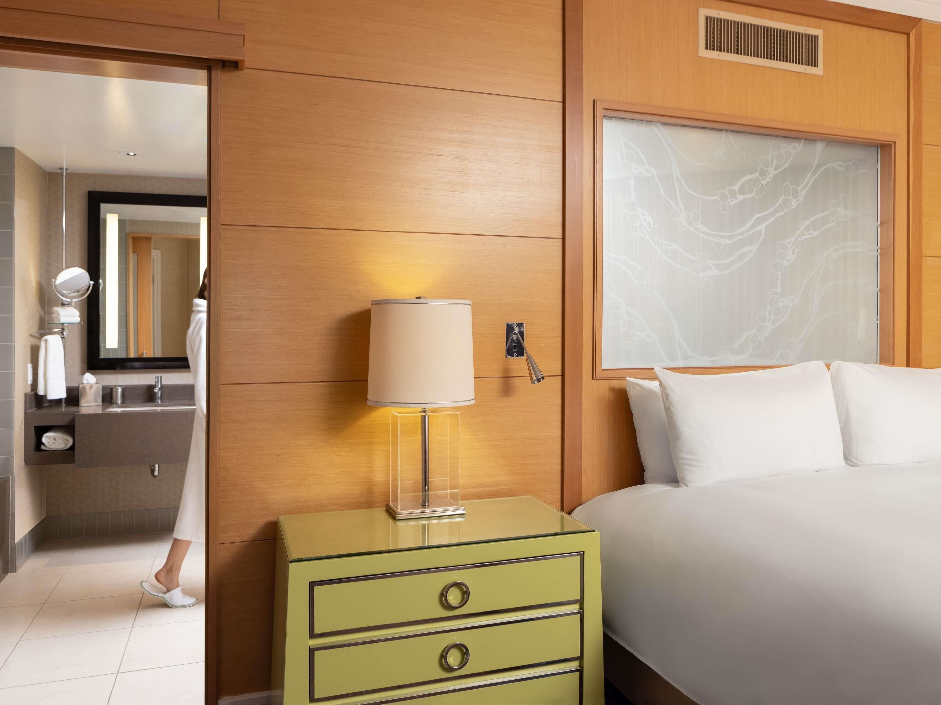 Hotelkamer met een bed, nachtkastje met een lamp, spiegel, badkamer met wastafel en handdoekhouder, en een houten wandpaneel.