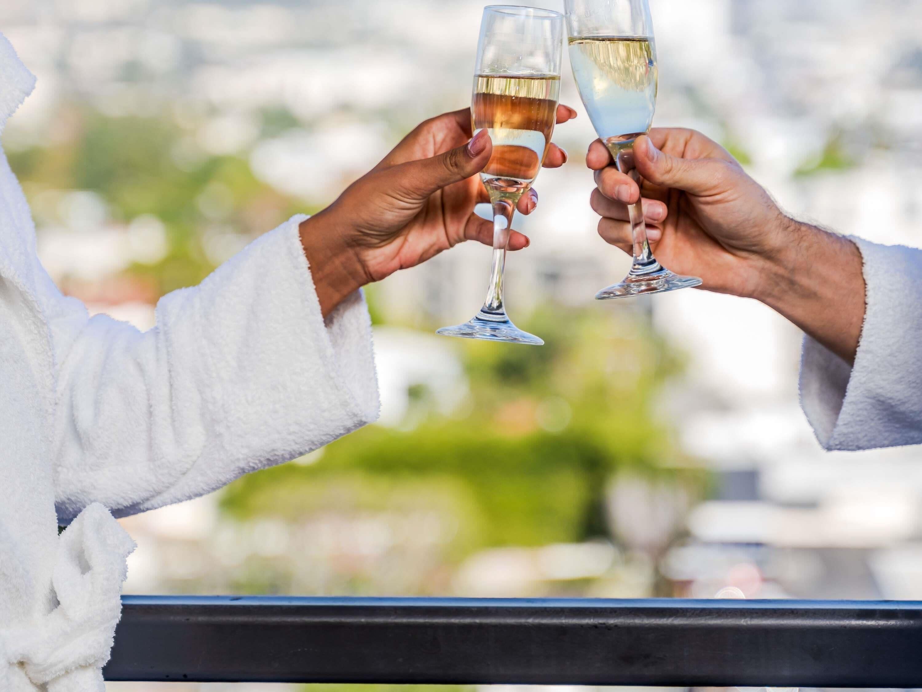 Twee personen proosten met champagneglazen, gekleed in witte gewaden, met een onscherpe achtergrond buiten.