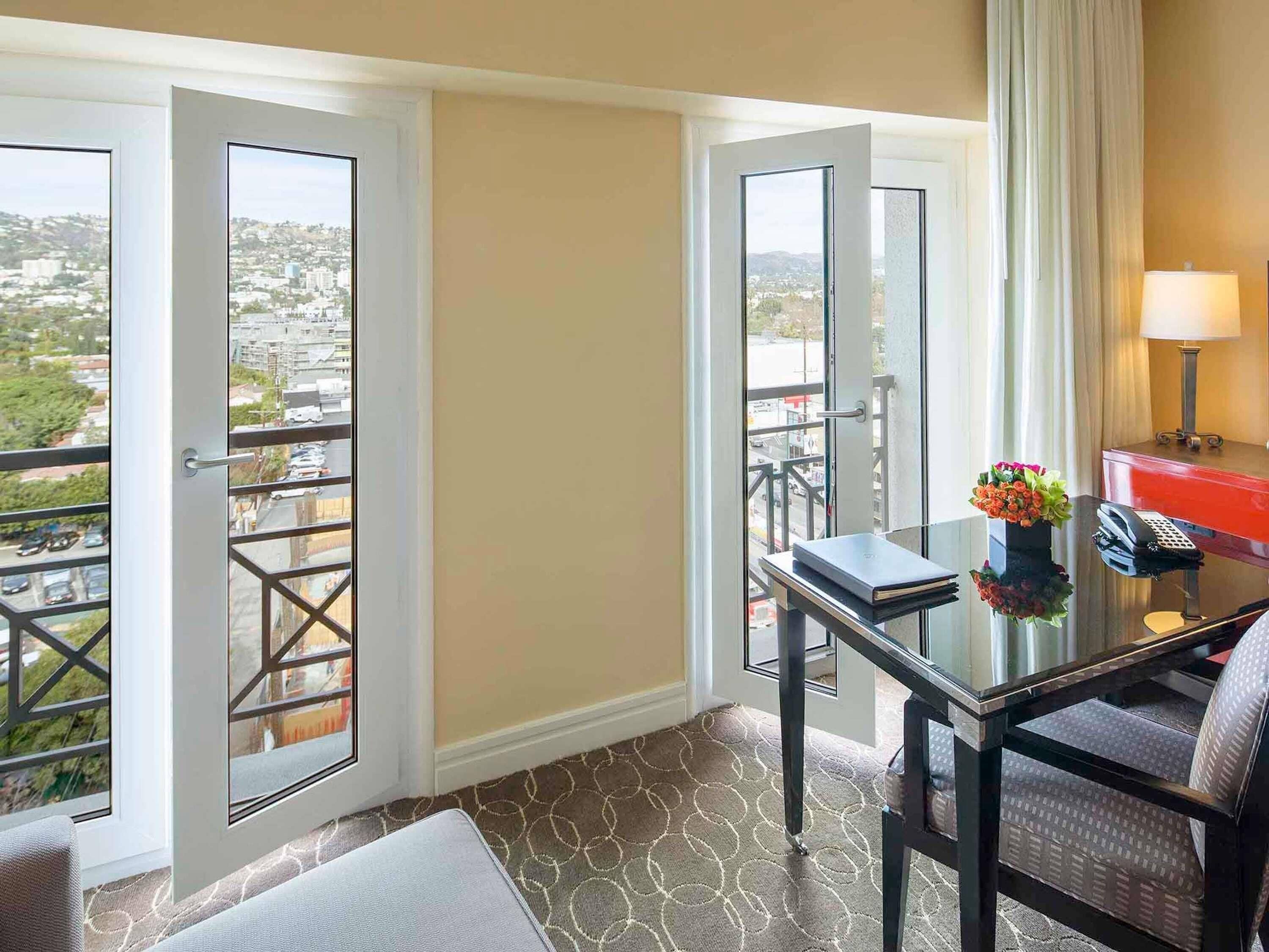 Habitación de hotel con balcón, escritorio con lámpara, una silla y vista a la ciudad.