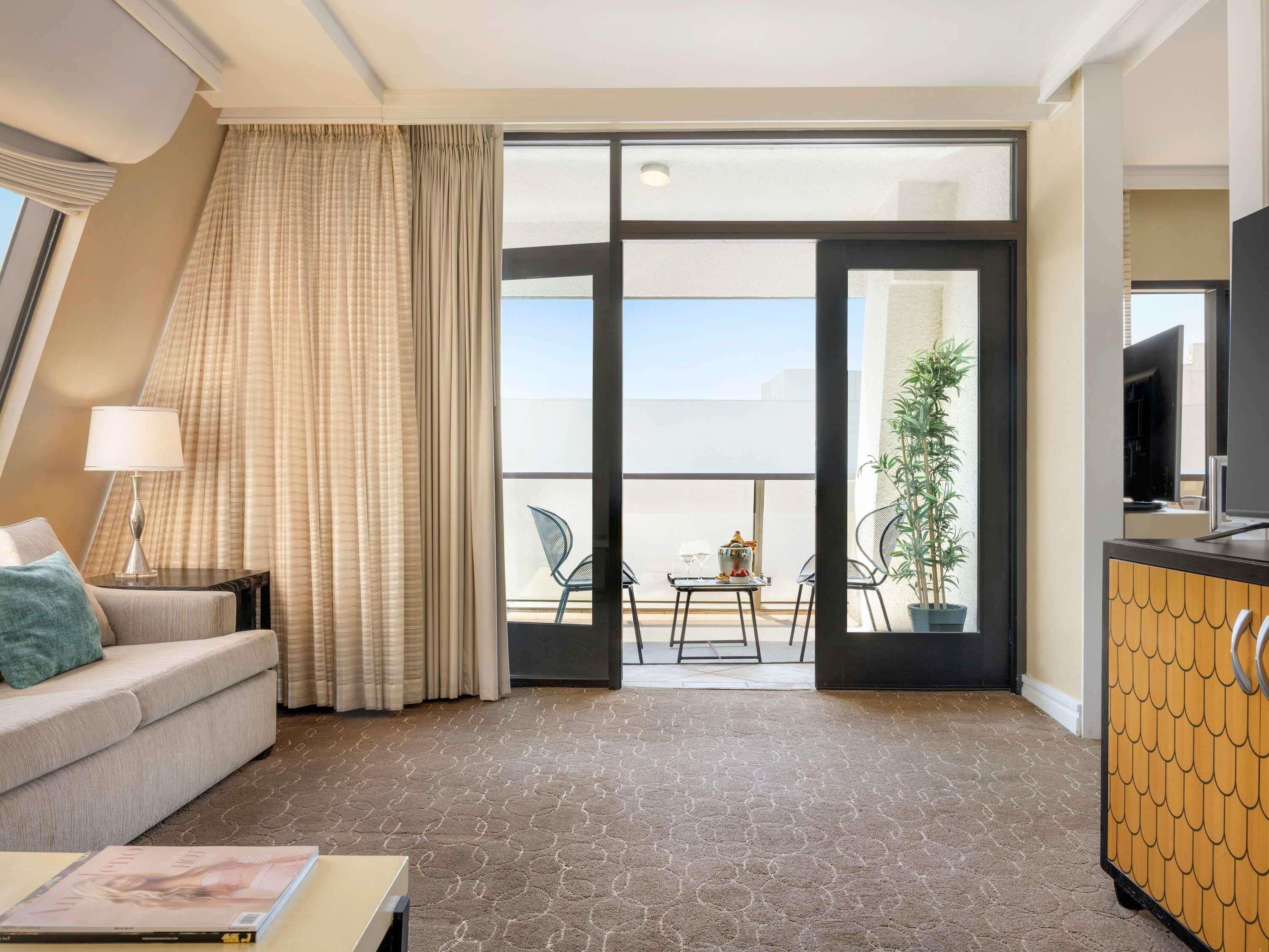 A hotel room with a sofa, a table, a lamp, a TV, a balcony with chairs, and a plant.