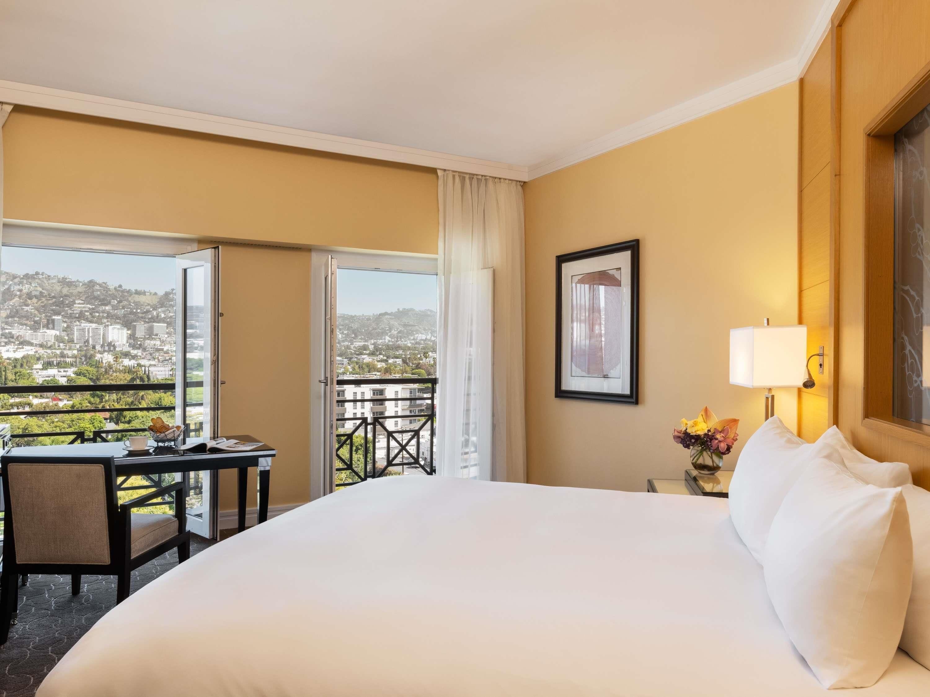 A hotel room with a large bed, a desk with a chair, a window with a view of a cityscape, and a framed picture on the wall.