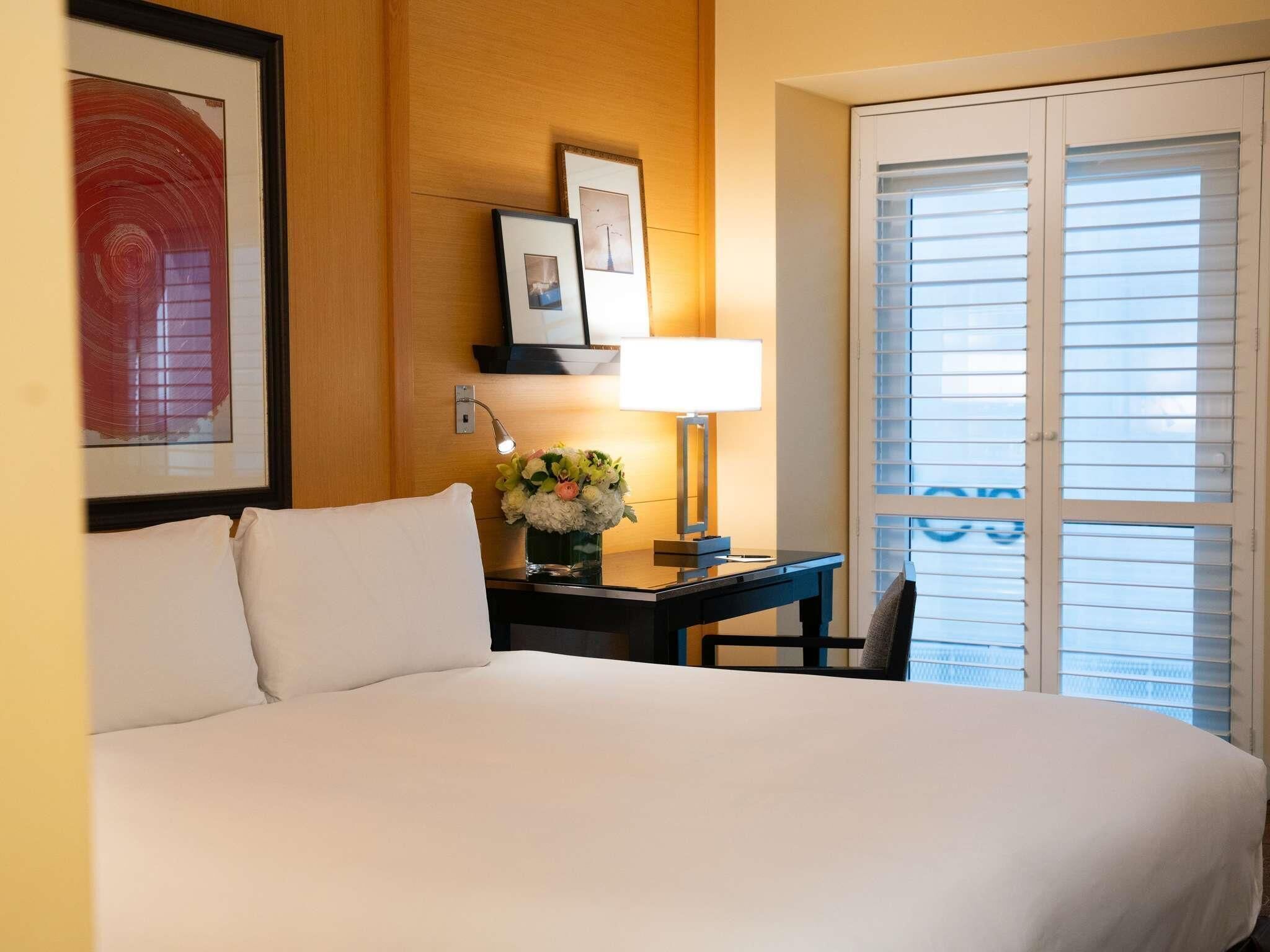Een hotelkamer met een bed, een bureau met lamp, een bloemetjesvaas en een raam met jaloezieën.