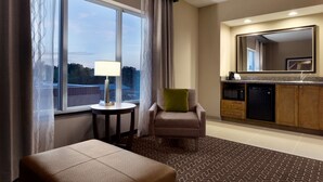 Ein Hotelzimmer mit großem Fenster, einem Sessel mit grünen Kissen, einem kleinen Tisch mit Lampe, einer Küchenzeile mit Spiegel und einem gemusterten Teppich.
