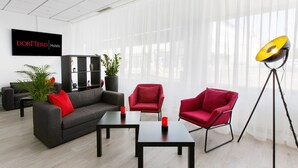 A modern hotel room with a grey sofa, red armchairs, a black coffee table, and a floor lamp. There is a bookshelf with plants and a TV mounted on the wall displaying 'DORMERO Hotels'. - Dormero Hotel Dessau-Rosslau (Dessau-Rosslau)
