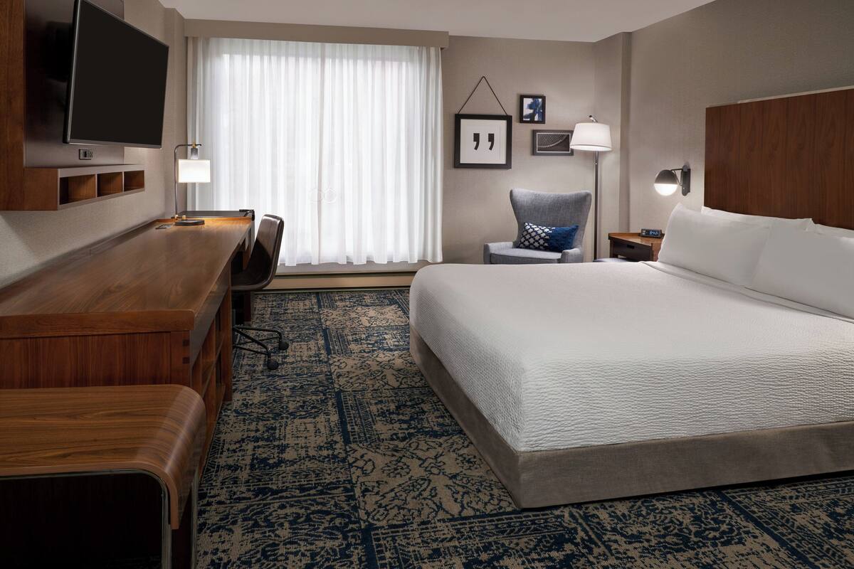 Une chambre d’hôtel avec un grand lit, un bureau avec une chaise, un téléviseur fixé au mur et une fenêtre avec des rideaux.