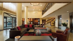 A spacious lobby with a sectional sofa, a coffee table, and a red and grey patterned rug. There is a staircase with a railing, an elevator, and a reception area with a counter and chairs.