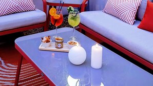 Un área de estar al aire libre y de estilo moderno con una mesa de vidrio, dos sillas acolchadas, una vela, un tarro y una bandeja con aperitivos.