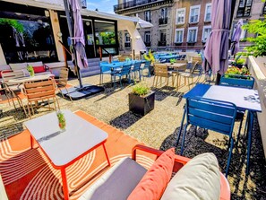 An outdoor seating area with tables and chairs, a small garden, and a building with large windows in the background. - Ibis Grenoble Centre Bastille (Grenoble)