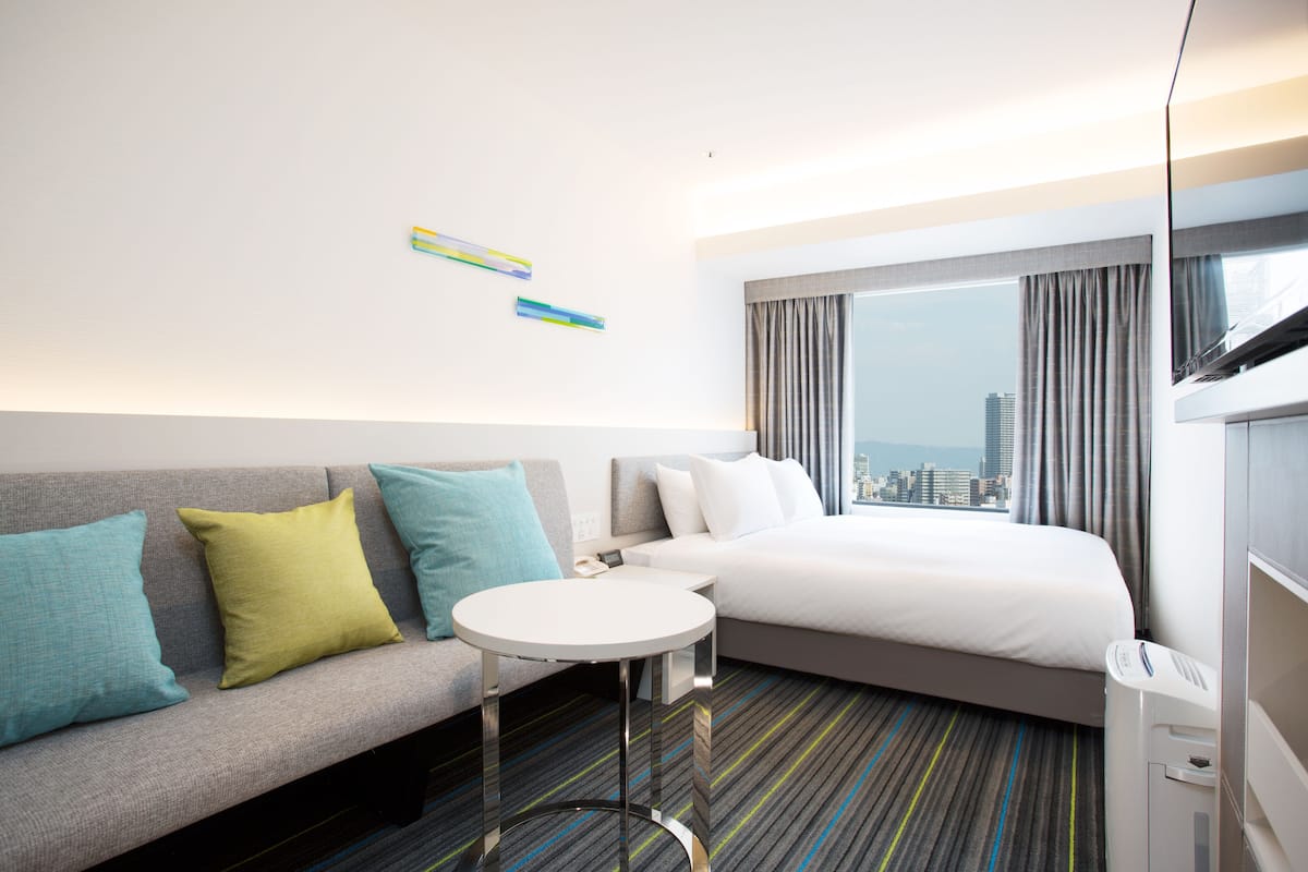 A modern hotel room with a grey sofa, a small round table, a bed with white linens, and a large window with curtains.