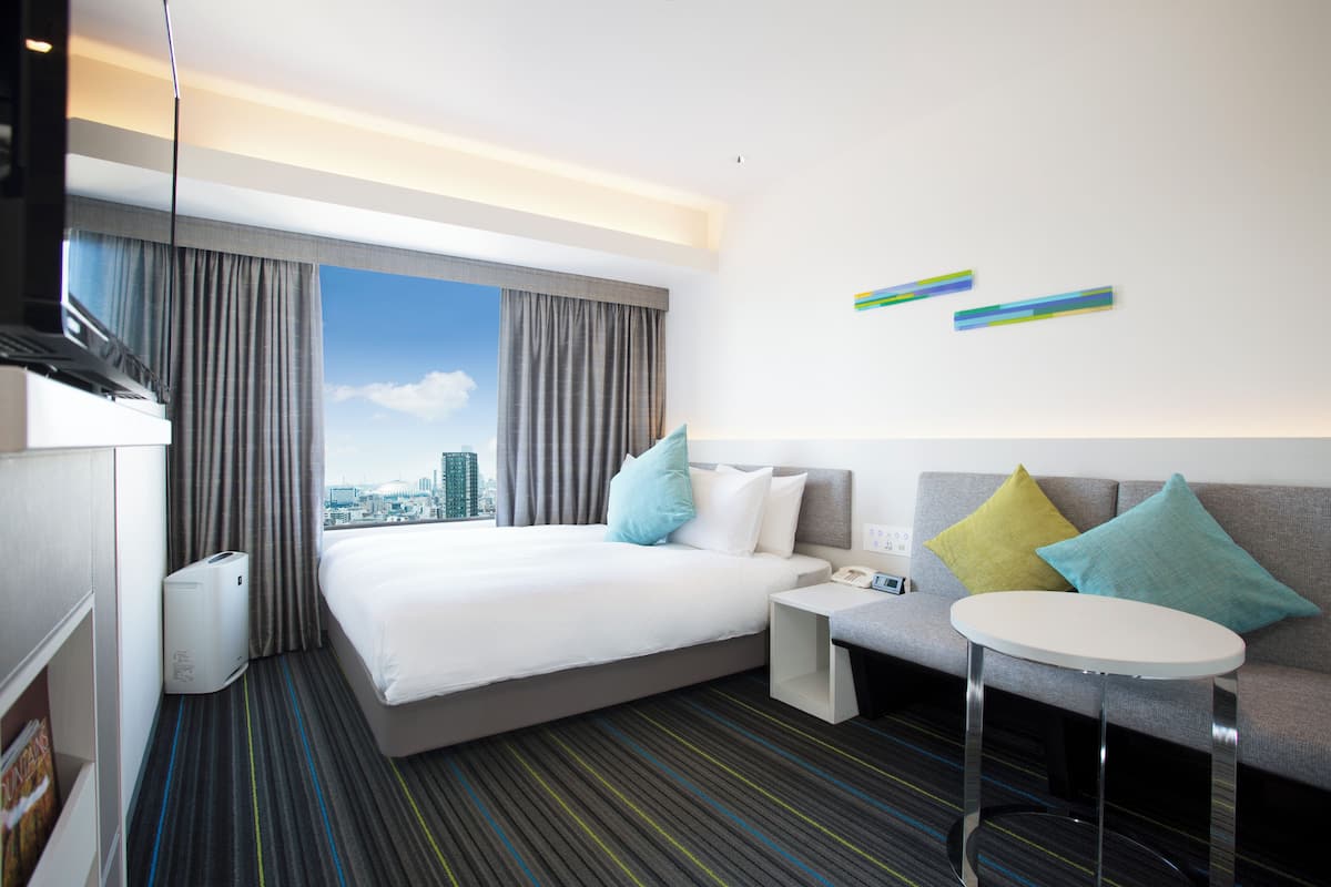 A hotel room with a large bed, a sofa, a small table, and a view of the city.