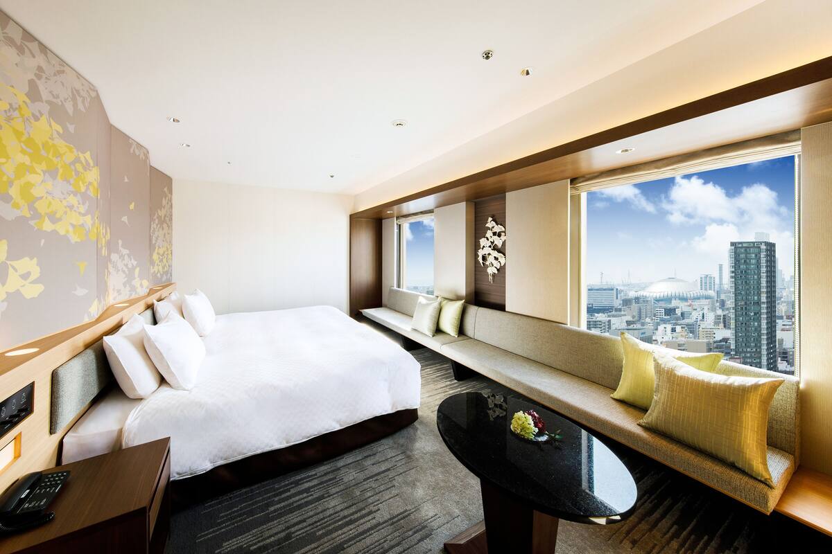 A modern hotel room with a large bed, a seating area with cushions, a desk with a phone, and a view of the city.