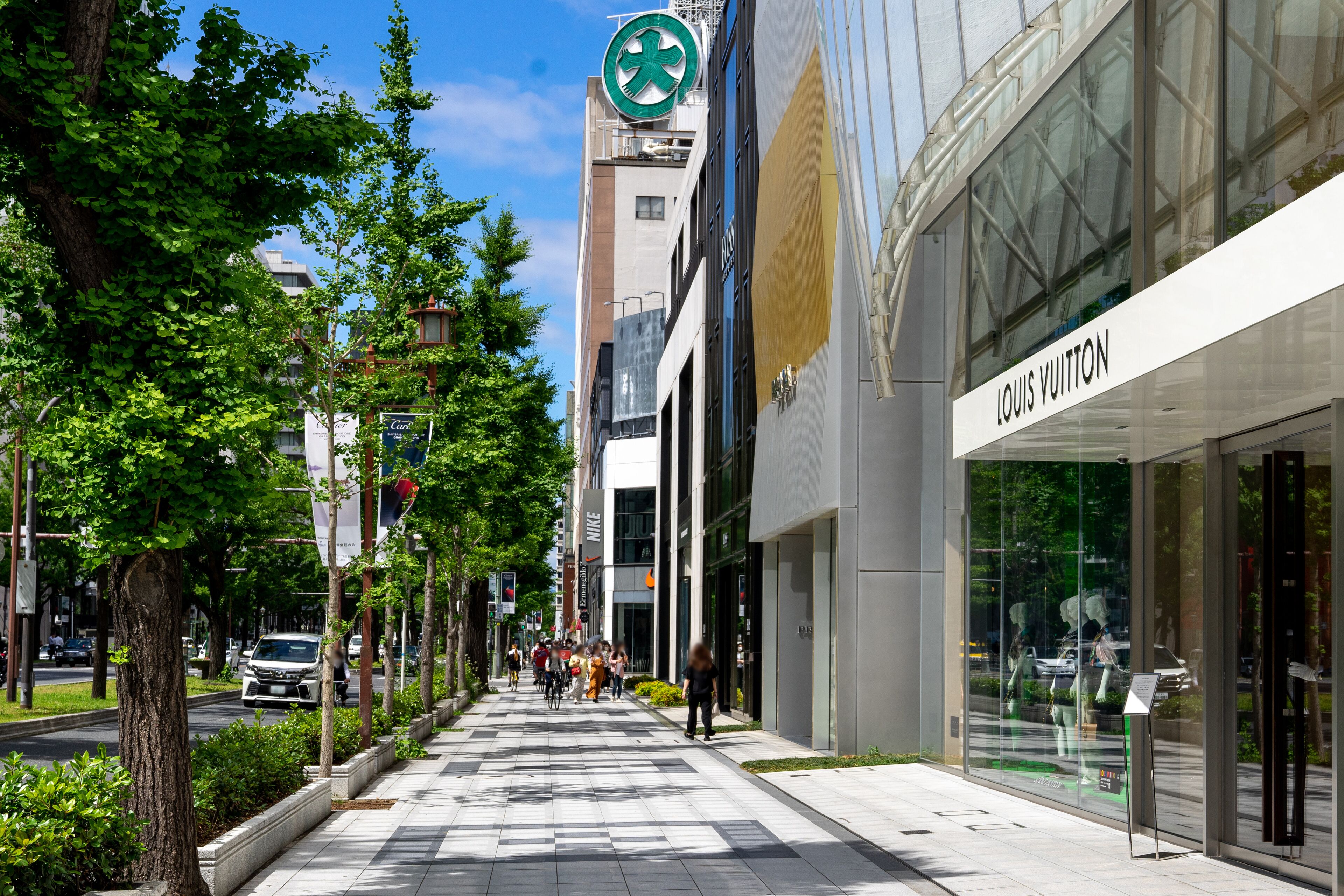 Una calle urbana moderna con una tienda Louis Vuitton, una farmacia y una acera bordeada de árboles.