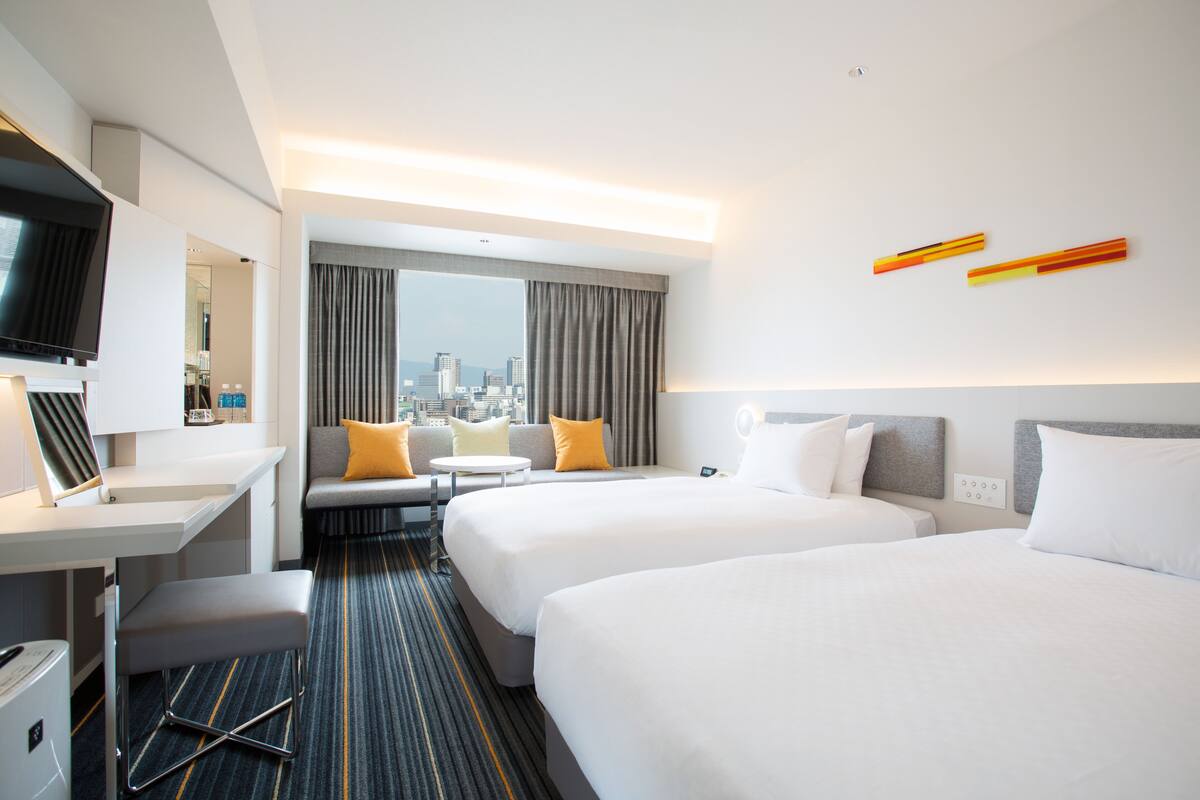 A hotel room with two beds, a desk, a TV, and a city view.