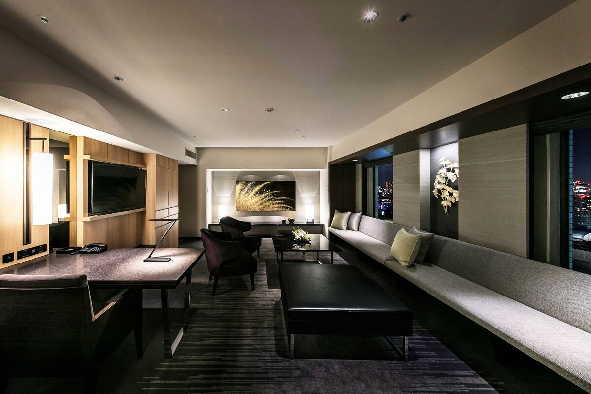 A modern hotel room with a large flat-screen TV, a sofa, a dining table with chairs, and a decorative wall piece.
