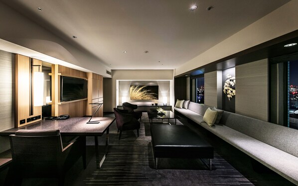 A modern hotel room with a large flat-screen TV, a sofa, a dining table with chairs, and a decorative wall piece.