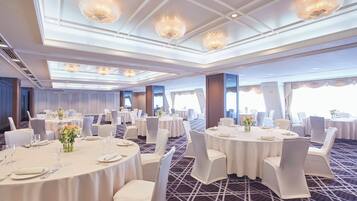 Uma sala de banquetes com mesas redondas, cadeiras brancas e lustres.