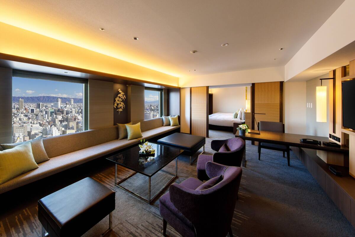 A modern hotel room with a city view, a sofa, a bed, a desk, and a TV.