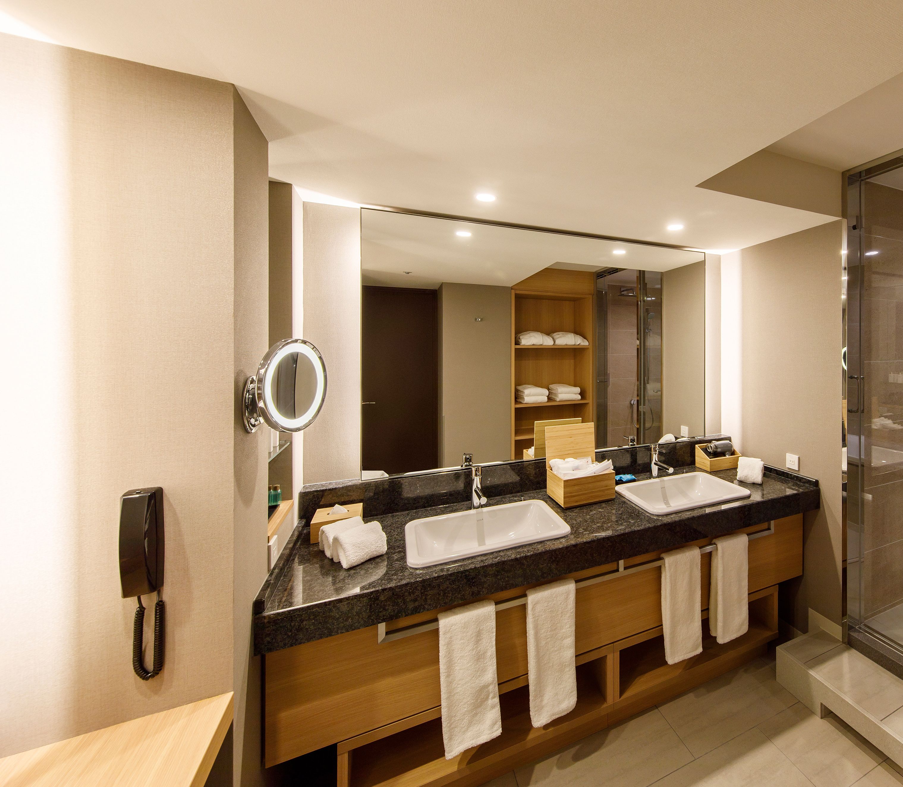 A hotel bathroom with a double sink vanity, a mirror, a glass shower enclosure, and a telephone.