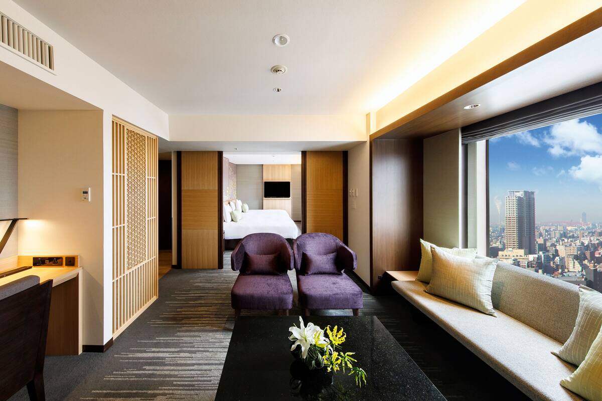 A modern hotel room with a bed, two purple armchairs, a sofa, a coffee table with flowers, and a city view.