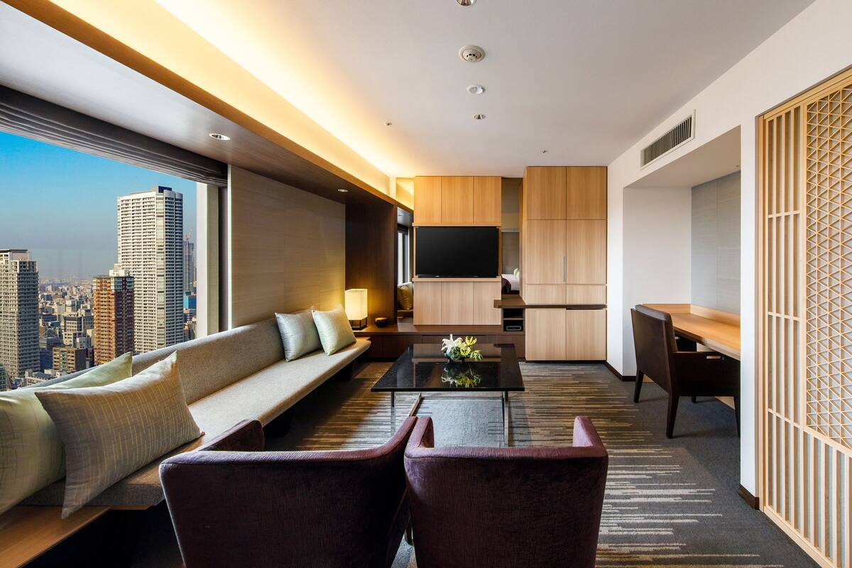 A modern hotel room with a large window, a sofa, a coffee table, a TV, and a desk.