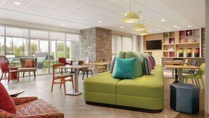 A spacious indoor area with a green sofa, wooden tables, and chairs, a stone wall, and a TV on a wooden shelf. - Home2 Suites by Hilton Williamsville Buffalo Airport (Buffalo)