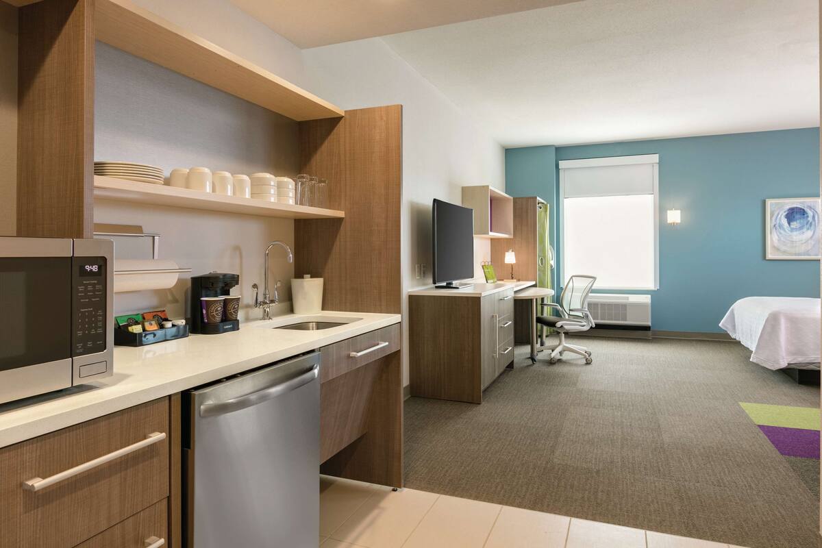 A modern hotel room with a kitchen area, a desk, and a bed.