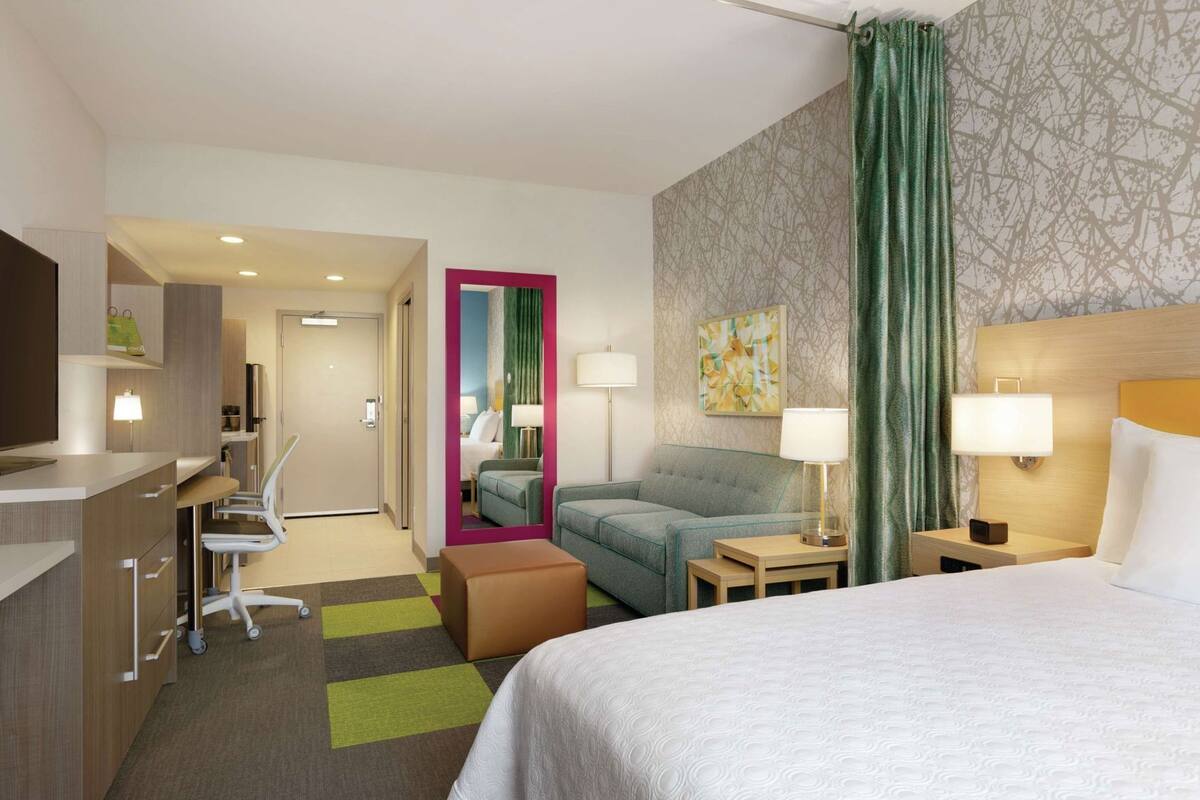 A hotel room with a bed, a desk, a chair, a sofa, a nightstand, a lamp, and a mirror.