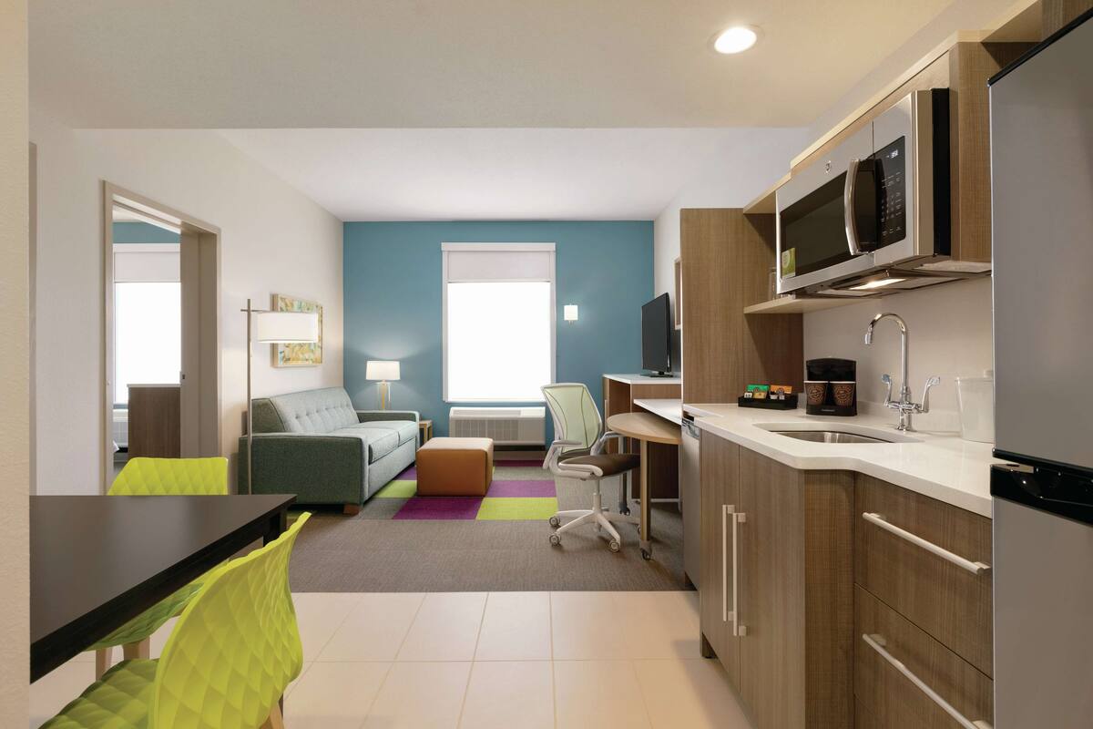 A modern kitchen with a microwave, refrigerator, and sink. There is a dining area with a table and chairs, and a living area with a sofa and a coffee machine.