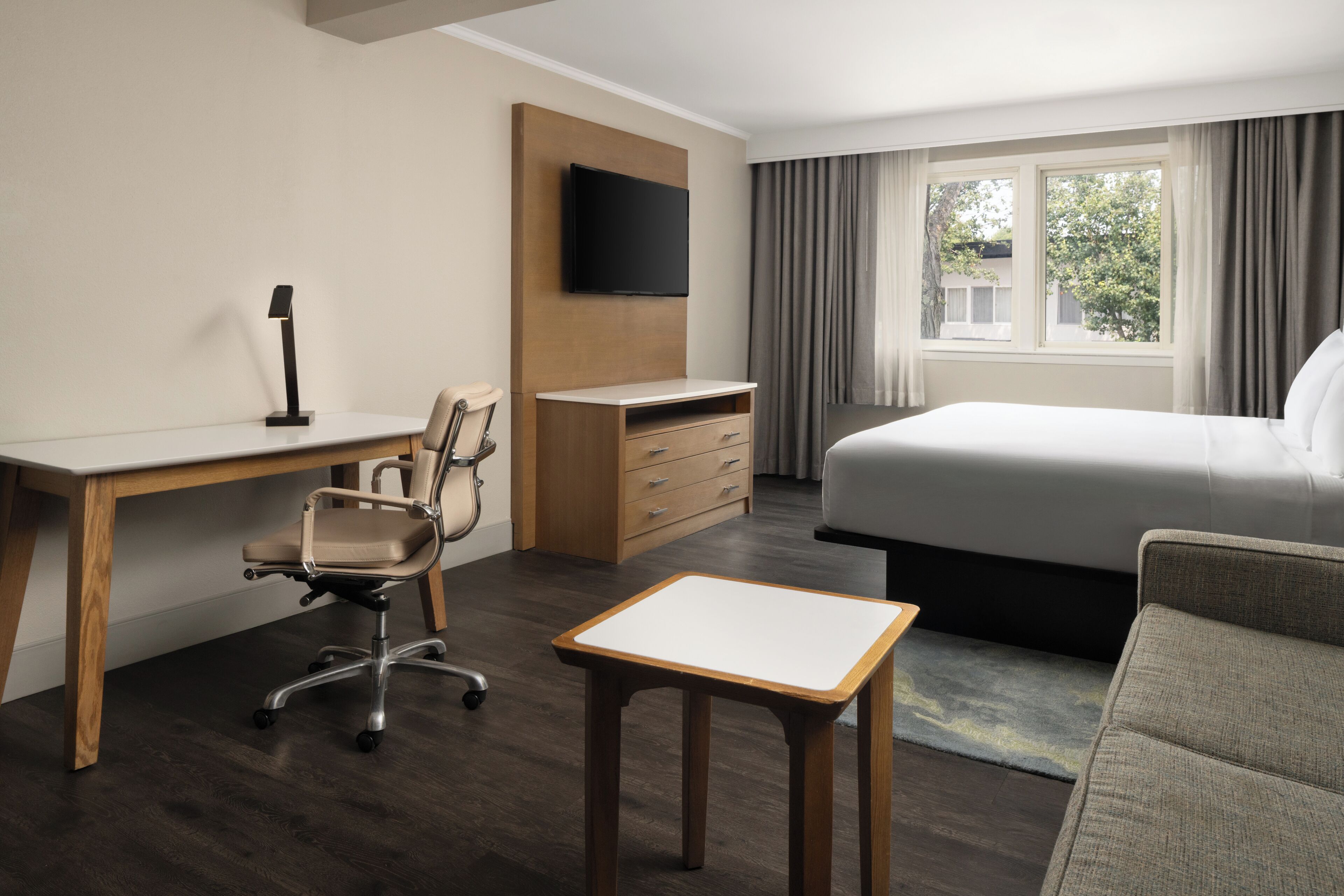 Habitación de hotel con cama, escritorio con silla, televisor en la pared y ventana con cortinas.