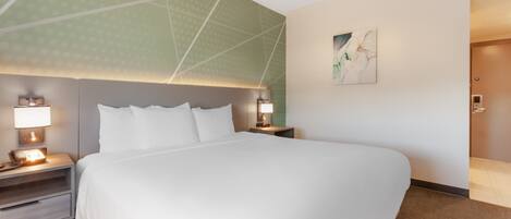 Une chambre d’hôtel avec un grand lit, deux tables de chevet avec des lampes, une œuvre d’art accrochée au mur et une porte donnant sur une autre pièce.