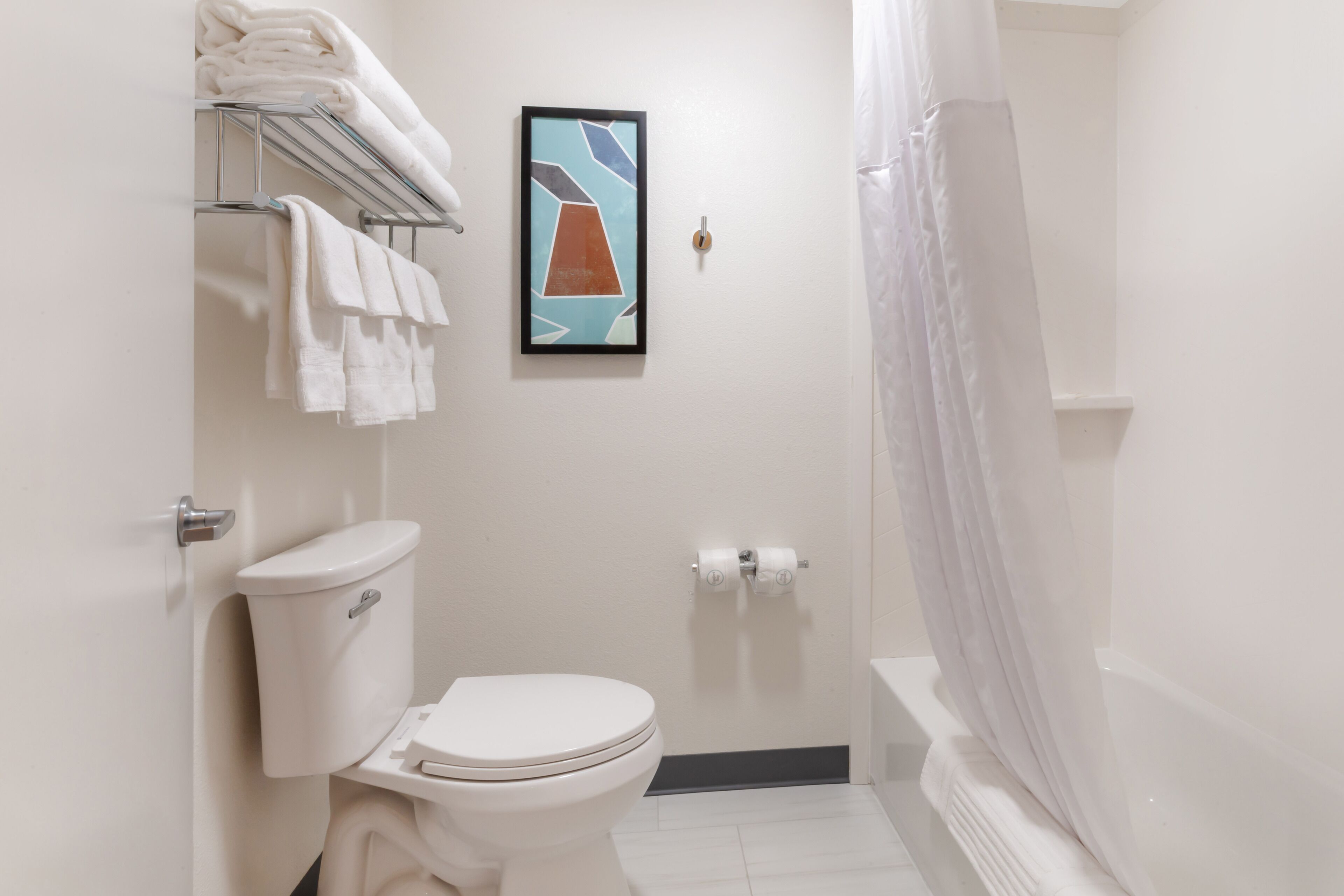 A bathroom with a toilet, a towel rack with folded towels, a framed picture on the wall, and a shower curtain.