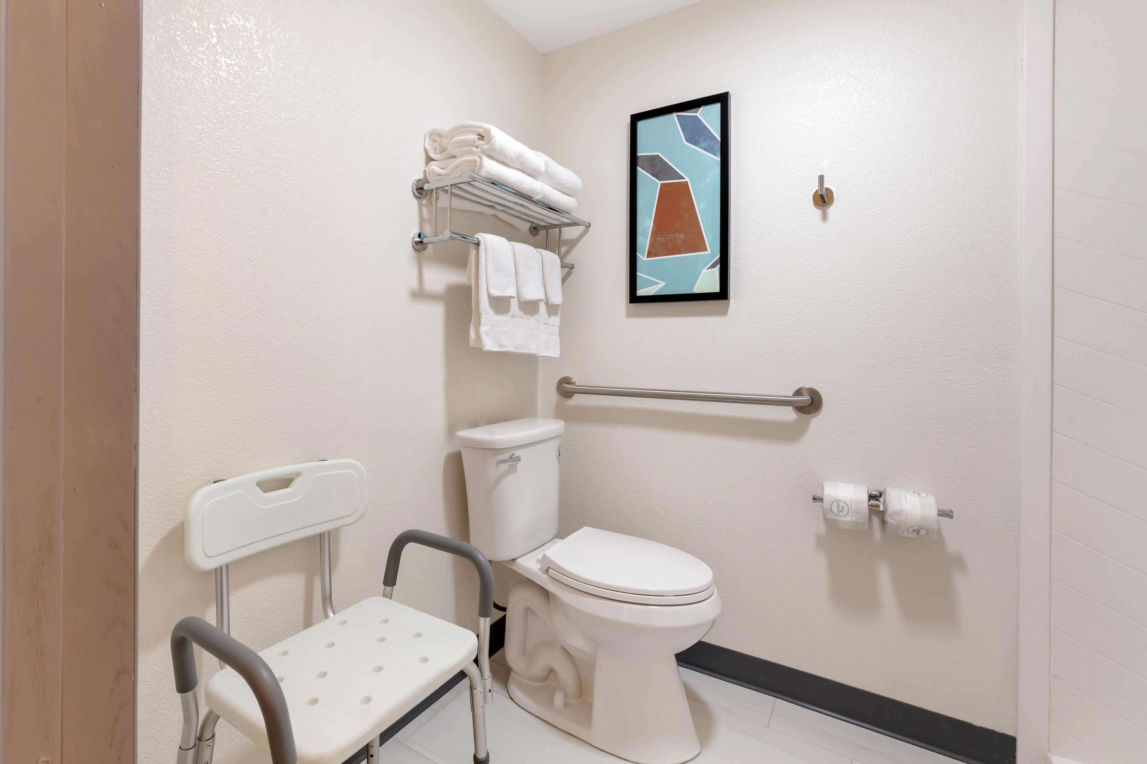 A bathroom with a white toilet, a handrail, a grab bar, a towel rack with folded towels, and a framed abstract art piece on the wall.