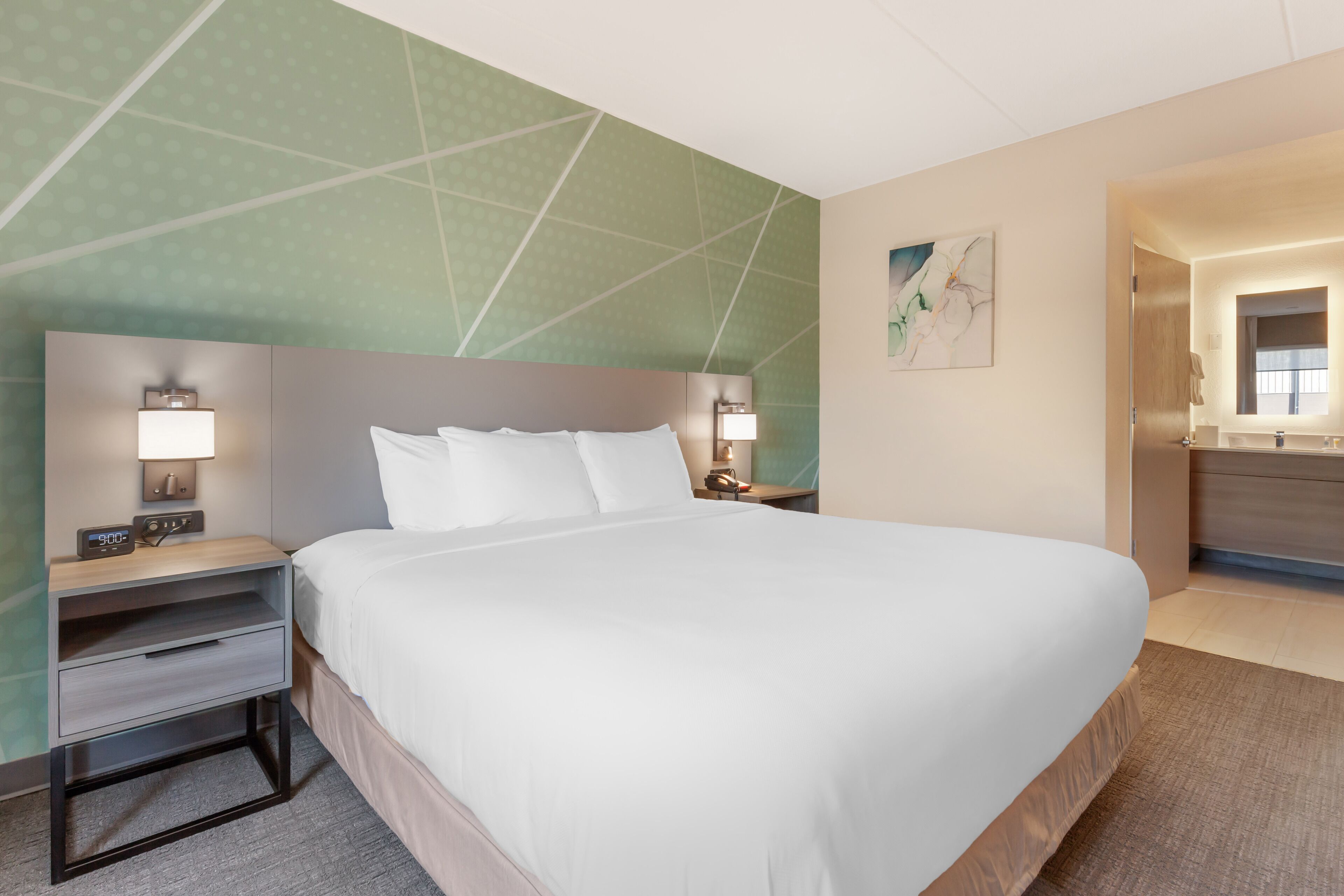 A hotel room with a large bed, bedside tables with lamps, a nightstand, a wall-mounted artwork, and a bathroom visible through an open door.