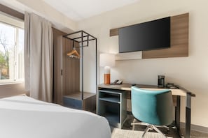 A hotel room with a bed, desk, chair, TV, and a hanging coat rack.