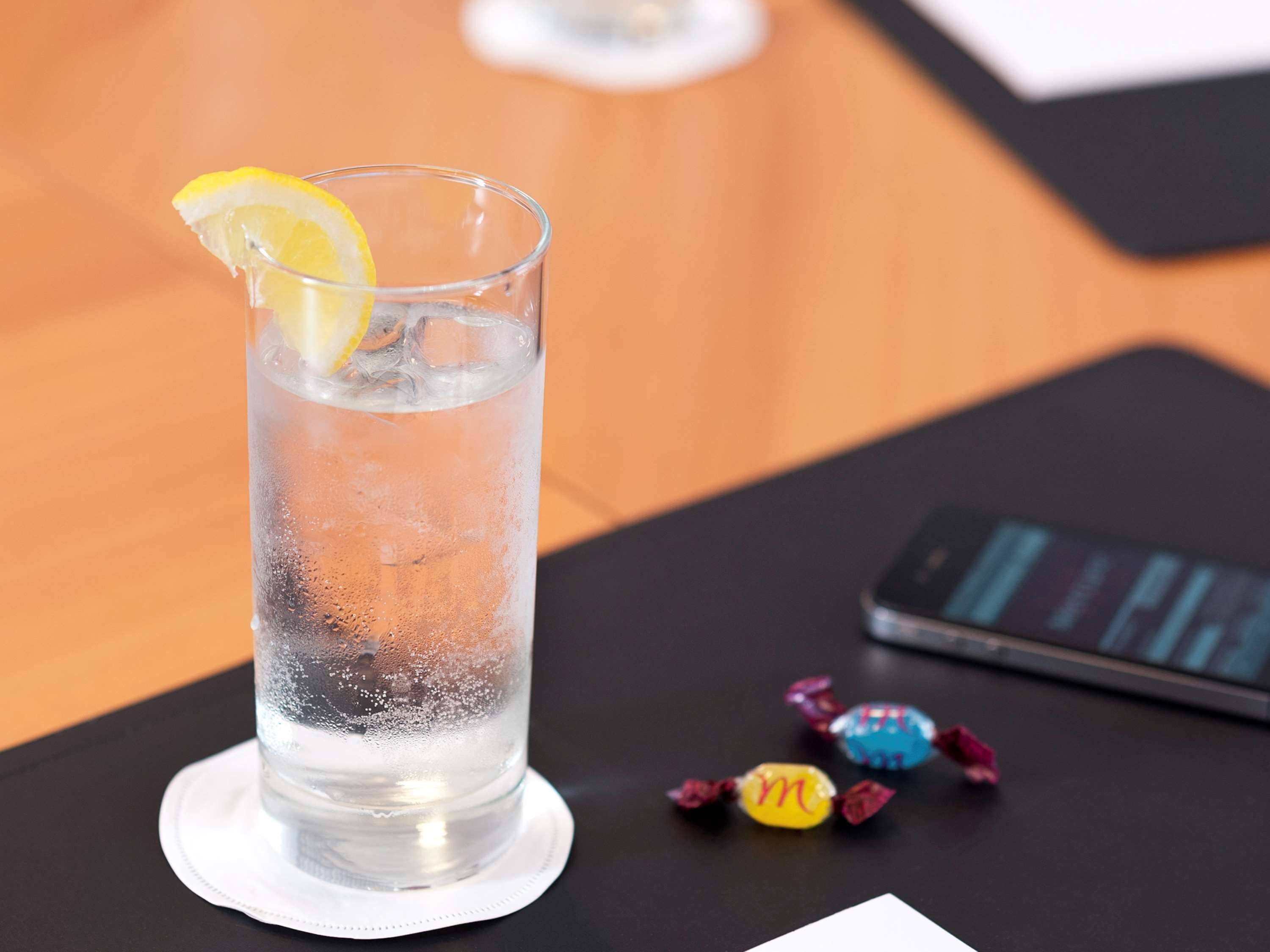 Un verre transparent contenant des glaçons et une tranche de citron, posé sur un dessous de verre, un téléphone intelligent et des bonbons colorés emballés individuellement, le tout sur une table.
