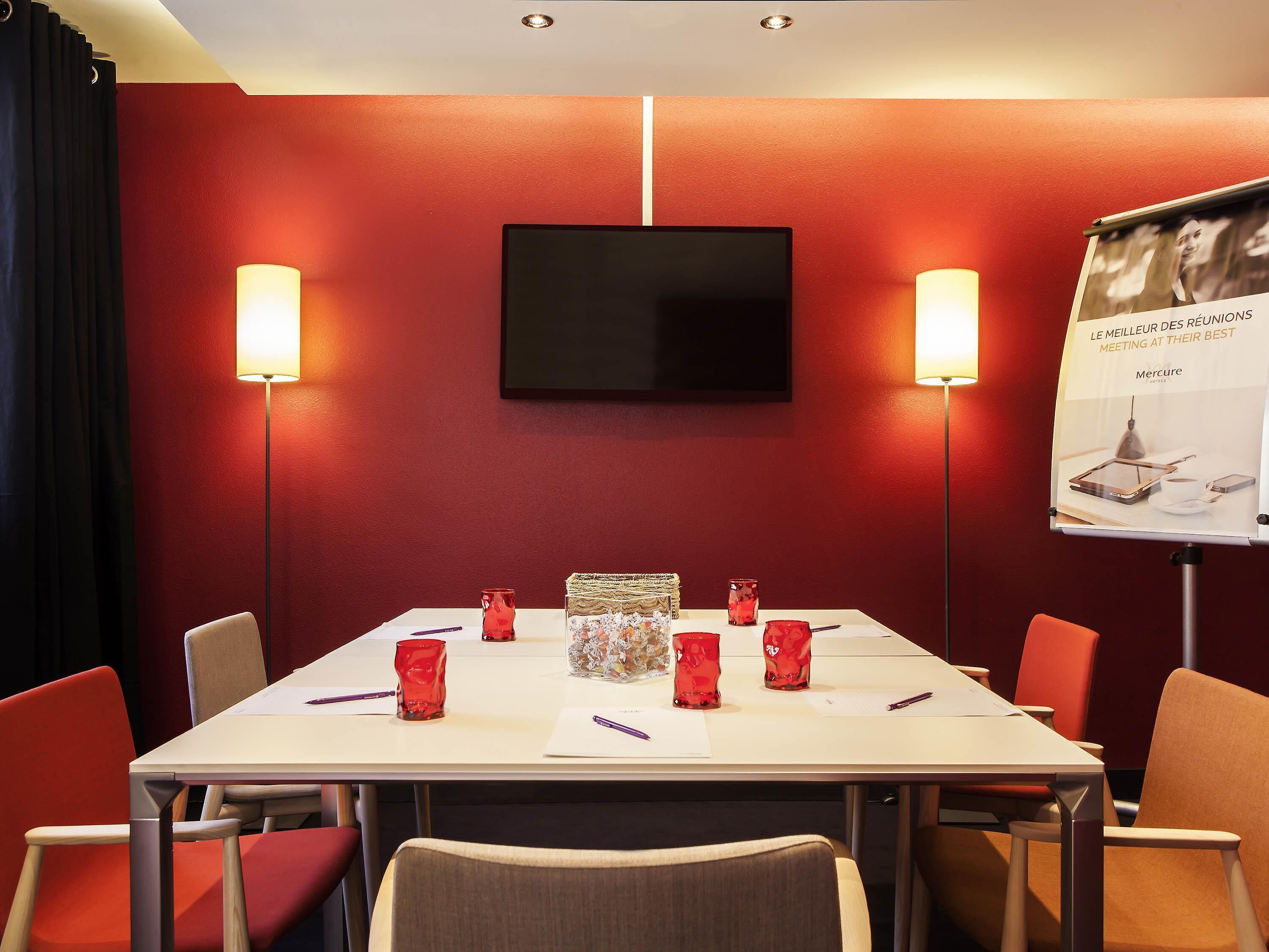 A conference room with a white table, red chairs, a flat-screen TV, and a poster on the wall.